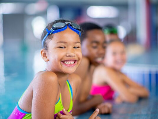 Girl smiling leaning out of pool with googles on head